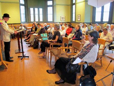 Gus at the podium reading with the audience at Westmount Library and Karin in foreground